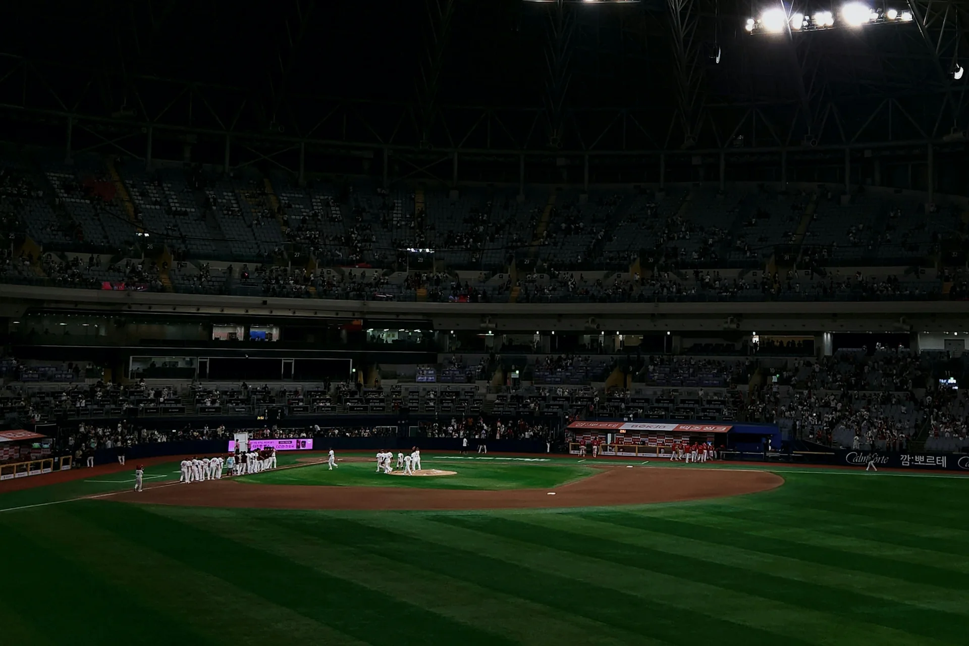 A night KBO baseball game underway in a packed South Korean stadium, floodlights blazing over the infield and players gathered near the mound